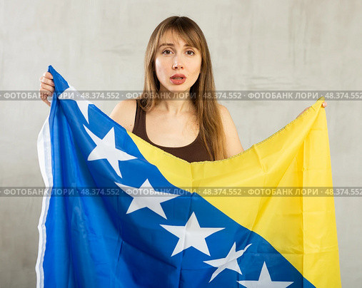 Sad young woman holding flag of Bosnia and Herzegovina against unicoloured background. Стоковое фото, фотограф Яков Филимонов / Фотобанк Лори