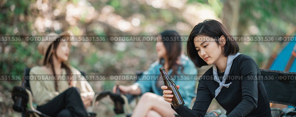Selective focuse, beautiful Asian woman and friends travelers  relaxing at front of camping tent, They enjoy to talking and drinking beer with fun and happy together. Стоковое фото, агентство Ingram Publishing / Фотобанк Лори