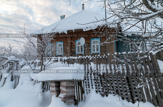 Село Домнино, Сусанинский район Костромской области. Зимний пейзаж. Морозно, медленно спускаются сумерки. Деревенский одноэтажный дом со спутниковой антеной за слегка покосившимся забором утонули в снегу (2022 год). Стоковое фото, фотограф Наталья Николаева / Фотобанк Лори