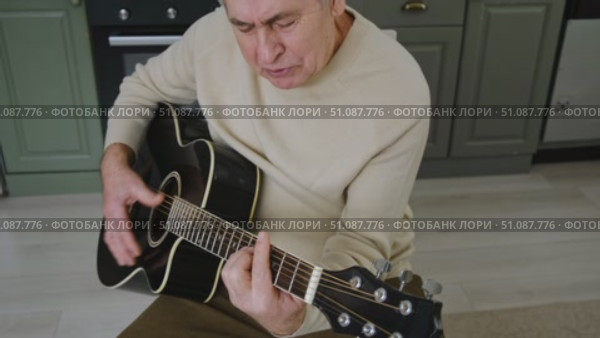 Senior man is playing guitar. Elderly man sitting on the sofa and playing guitar. Portrait of a gray-haired mature man in a sweater and glasses learning to play. Enjoying retirement life at home. Стоковое видео, видеограф Марина Демидюк / Фотобанк Лори