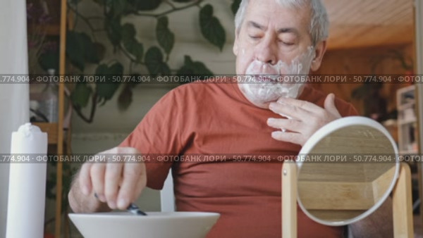 Senior Man shaving his face. Old male shaves beard and mustache with shaver. Elderly gray haired man looking at the mirror, applies shaving cream foam and using razor. Skin hygiene. Daily body care. Стоковое видео, видеограф Марина Демидюк / Фотобанк Лори