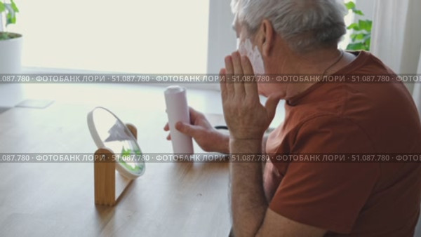 Senior Man shaving his face. Old male shaves beard and mustache with shaver. Elderly gray haired man looking at the mirror, applies shaving cream foam and using razor. Skin hygiene. Daily body care. Стоковое видео, видеограф Марина Демидюк / Фотобанк Лори