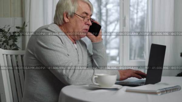 Serious senior man wearing glasses Talking mobile phone sitting at desk at home workplace with laptop. Retired male working chatting colleagues. Old worker talk on cellphone work on computer gadget. Стоковое видео, видеограф Марина Демидюк / Фотобанк Лори