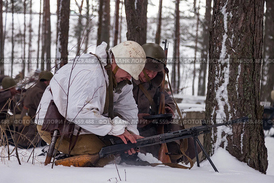 Sestroretsk, Russia - February 19, 2023: Two soldiers huddle in the snow, repairing their weapon as winter's chill descends, surrounded by tall trees and signs of battle. Редакционное фото, фотограф Aleksandr Golubev / Фотобанк Лори
