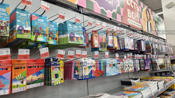 Shelves in a supermarket with a wide range of colored pencils and art supplies. The display case features sets from various brands: Krasin, Kruzhok and Dilin, Erich Krause, and other manufacturers. The pencils are packed in cardboard boxes with bright designs - images of animals, balloons, landscapes, cartoon characters, and geometric patterns. The range includes sets of 6, 12, 18, 24, and more colors. Prices are posted on individual price tags with promotional discounts. At the bottom of the shelf, stacks of sketchbooks and notebooks are visible. The photo is clear, with good detail, ideal for online stationery stores, catalogs of school supplies, educational materials, and advertising banners for stores specializing in children's creativity and preparation for school. Редакционное фото, фотограф Анна Кривицкая / Фотобанк Лори