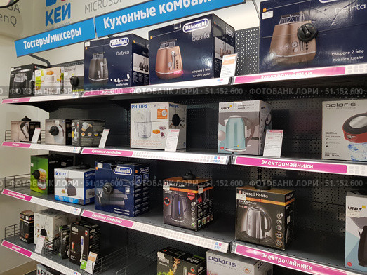 Shelves of a home appliance store with a variety of kitchen appliances. The display case features electric kettles, toasters, food processors, blenders and other kitchen appliances. Boxes with goods are located on metal shelves with price tags and information signs. Among the brands you can see Delonghi, Philips, Polaris, Vitek, Unit, Russell Hobbs, Braun. The top shelves are occupied by toasters, below are food processors, and a separate row is dedicated to electric kettles. The photo conveys the atmosphere of a modern electronics store and demonstrates the range of goods for cooking and household convenience. (2019 год). Редакционное фото, фотограф Анна Кривицкая / Фотобанк Лори