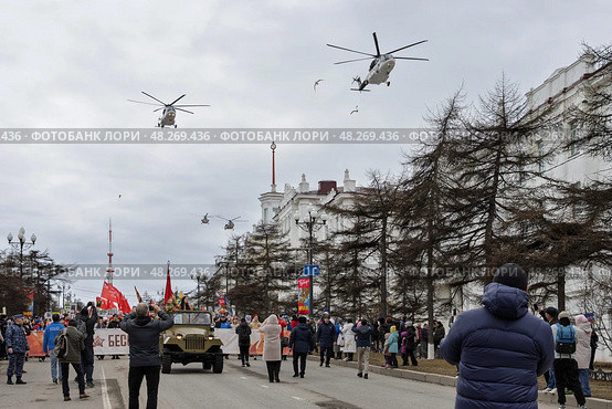 Шествие «Бессмертного полка» в Магадане, 9 мая 2025 года. День Победы. По проспекту Ленина идёт колонна демонстрантов с транспарантом «Бессмертный полк». Возглавляет колонну старый советский автомобиль ГАЗ. Участники демонстрации несут красные флаги и портреты ветеранов войны. Над городом летят вертолёты. Празднование 80-летия победы Красной армии и советского народа над нацистской Германией в Великой Отечественной войне 1941–1945 годов. Город Магадан, Магаданская область, Россия. Редакционное фото, фотограф Андрей Степанов / Фотобанк Лори