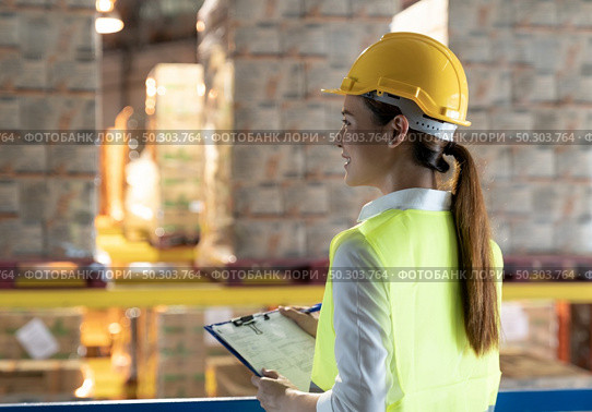 Side view of asian female warehouse worker with archboard flip chart look inventory with dustrubution warehouse in background. Reopening business warehouse technology and logistic concept. Стоковое фото, агентство Ingram Publishing / Фотобанк Лори