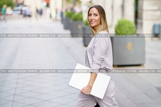 Side view of cheerful adult businesswoman with long blond hair in stylish outfit smiling, and looking away while strolling on paved street with laptop in hand before work. Happy female manager with laptop walking on street before meeting. Стоковое фото, агентство Ingram Publishing / Фотобанк Лори