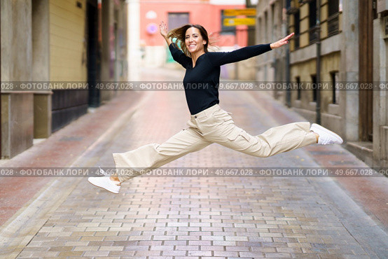 Side view of delighted young female with brown hair in casual clothes and sneakers smiling and looking at camera while jumping on narrow paved street. Excited young woman jumping on paved street and smiling. Стоковое фото, агентство Ingram Publishing / Фотобанк Лори