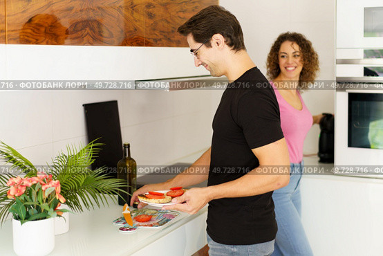 Side view of happy adult male looking down while standing with smiling conversing female at counter, with wine bottle flower pot and preparing healthy salad in kitchen at home in daylight. Happy adult man standing near cooking range with woman and arranging salad in kitchen. Стоковое фото, агентство Ingram Publishing / Фотобанк Лори