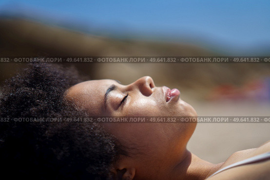 Side view of peaceful black female tourist with Afro dark hair, relaxing on sandy beach with closed eyes, and sunbathing on sunny day during summer holiday. Young African American woman lying on beach with closed eyes. Стоковое фото, агентство Ingram Publishing / Фотобанк Лори