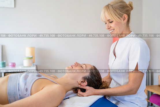 Side view of smiling female blond hair physiotherapist rubbing neck of client lying face up in bed and closing eyes during massage treatment. Cheerful female osteopath treating patient in hospital. Стоковое фото, агентство Ingram Publishing / Фотобанк Лори