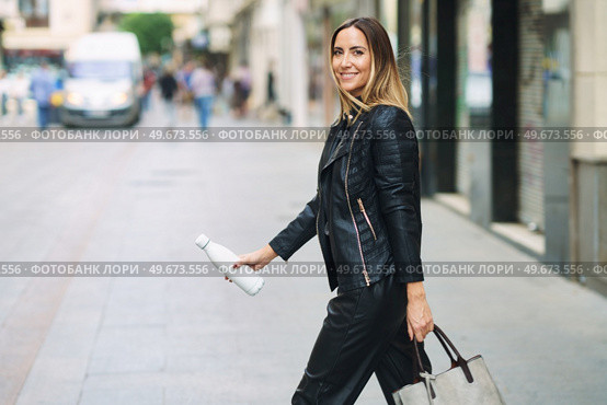 Side view of stylish female in leather pants and jacket carrying bag and takeaway reusable bottle while commuting on town street looking at camera. Smart casual lady with bag and bottle walking on street. Стоковое фото, агентство Ingram Publishing / Фотобанк Лори