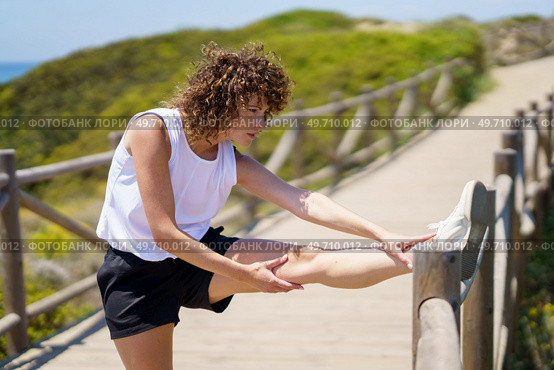 Side view of young female with curly hair in sportswear looking away while standing with stretched leg on bridge fence and soothing with hands in sunlight against blurred nature. Young sportswoman stretching leg on wooden bridge. Стоковое фото, агентство Ingram Publishing / Фотобанк Лори