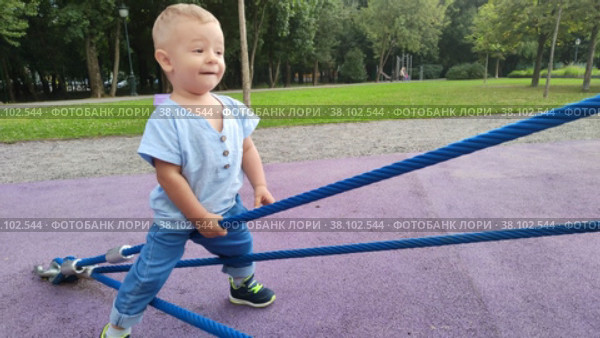 Симпатичный малыш со светлыми волосами играет на площадке в городском парке. Мальчик играет с толстыми веревками и лазает по ним. Счастливое детство. Ребенок учится ходить. Уличная площадка детского сада.  Cute blonde baby boy is playing on the playground in the city park. The child plays with thick ropes and climbs on them. Happy childhood. Kid is learning to walk. Outdoor playground for kindergarten. Стоковое видео, видеограф Ирина / Фотобанк Лори