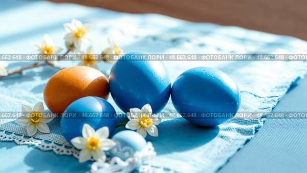 Синие и оранжевые крашенные яйца лежат на голубой скатерти. Blue and orange colored eggs are lying on a blue tablecloth. Стоковое AI-изображение, AI-художник Смирнова Светлана / Фотобанк Лори