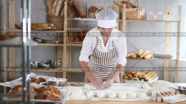 Skilled female baker in striped apron carefully handling individual dough portions at flour-covered worktable, surrounded by freshly baked bread, ingredients and tools in artisan bakery. Стоковое видео, видеограф Яков Филимонов / Фотобанк Лори