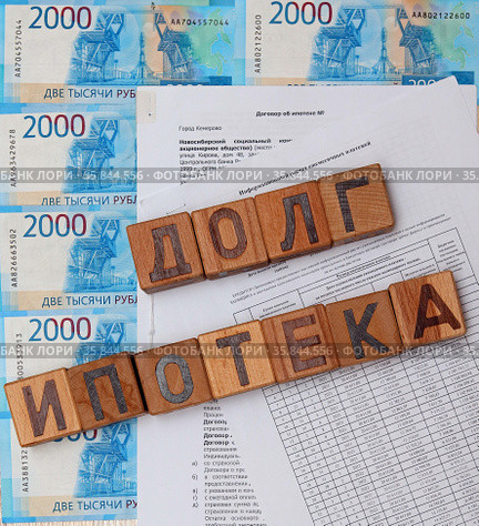 Слова "долг", "ипотека", сложенные из деревянных кубиков с буквами на фоне из российских денежных купюр, договора по ипотеке и графика платежей (2021 год). Редакционное фото, фотограф Цибаев Алексей / Фотобанк Лори
