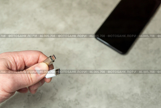 Смартфон на сером фоне и два кабеля USB-micro и USB Type-C в мужской руке на сером фоне. Кабель для заряда бытовой электроники. Редакционное фото, фотограф Sergei Gorin / Фотобанк Лори