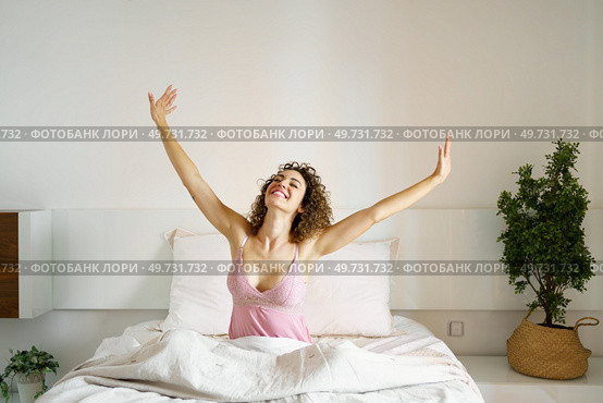 Smiling adult female with curly hair and with eyes closed, while sitting on cozy bed in daylight and stretching arms sideways with open palms in bedroom with green plant. Cheerful woman sitting on bed in sleepwear. Стоковое фото, агентство Ingram Publishing / Фотобанк Лори