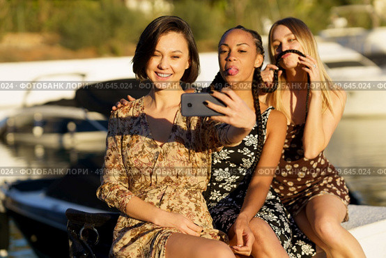 Smiling ethnic woman taking self portrait on cellphone with black girlfriend showing tongue and blonde making grimace on pier. Funny diverse women taking selfie on smartphone on seafront. Стоковое фото, агентство Ingram Publishing / Фотобанк Лори