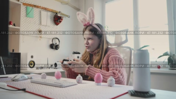 Smiling girl in bunny ears and futuristic glasses enjoys gaming, gripping controller with excitement. Immersed in virtual adventure, celebrates success in digital world, radiating joy and enthusiasm. Стоковое видео, видеограф Енваев Андрей / Фотобанк Лори
