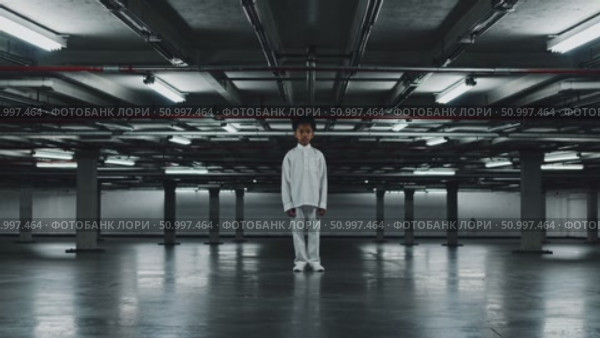 Solitary child wearing white, positioned centrally within empty concrete parking structure, industrial lighting casting stark shadows, expressing profound emotional isolation. Стоковое AI-видео, аниматор Данил Руденко / Фотобанк Лори