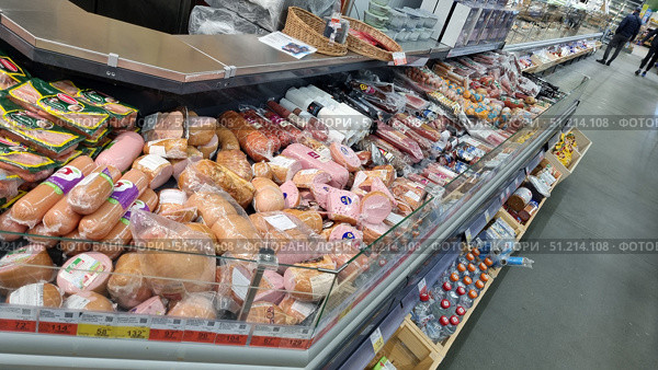 Supermarket counter with a large selection of sausages. The display case is filled with a variety of products: cooked sausages, smoked sausages, hot dogs, ham and cold meats in vacuum packaging. The products are neatly laid out in rows, many of them are packed in transparent film with labels. In the upper part of the display case, baskets and additional products are visible, and on both sides is a continuation of the meat department with a wide range of products. The lower shelves contain related products and drinks. The atmosphere of the photo reflects the reality of the supermarket - abundance and choice for customers. Редакционное фото, фотограф Анна Кривицкая / Фотобанк Лори