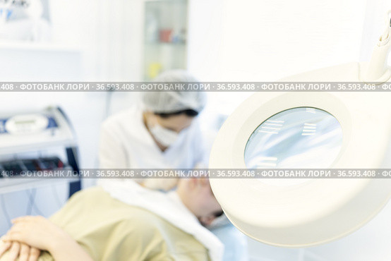 The doctor performs a cosmetic procedure against magnifying magnifying glass. The dermatologist's office with patients. Blurred background with cosmetic procedure for facial beauty skin. Cleansing. Стоковое фото, фотограф Ирина Ткачук / Фотобанк Лори