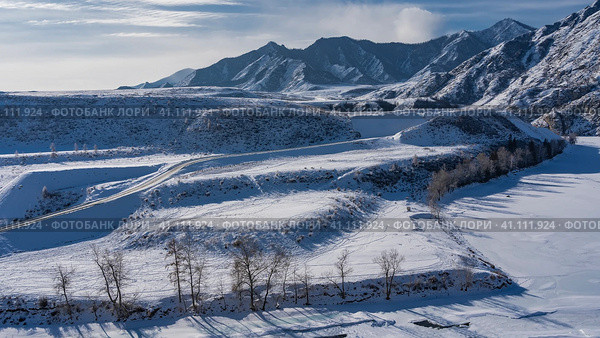 The frozen riverbed meanders. Bare trees along the shore. The road passes through a snow-covered valley. A picturesque mountain range against a background of blue sky and clouds. Altai. Стоковое фото, фотограф Вера Ивановна Кубарева / Фотобанк Лори