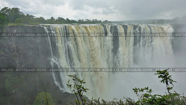 The magnificent Victoria Falls. The Zambezi River flows from the edge of the plateau into the gorge. Fog over powerful streams of water. There is green vegetation in the foreground. Zimbabwe (2020 год). Стоковое фото, фотограф Вера Ивановна Кубарева / Фотобанк Лори