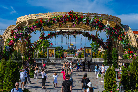 The photo was taken in the cultural and historical complex "New Chersonesus" in Sevastopol. In the foreground is an arched structure decorated with fresh flowers with hanging lamps, forming the entrance to the ceremonial site. Inside are icons, marble pedestals, a landscaped area and many visitors, including children and adults. In the distance, along the axis of the composition, is a majestic temple with a golden dome, symbolizing the spiritual axis of space. The atmosphere is bright, festive, with elements of traditional Orthodox culture and modern landscape design. Редакционное фото, фотограф Анна Кривицкая / Фотобанк Лори