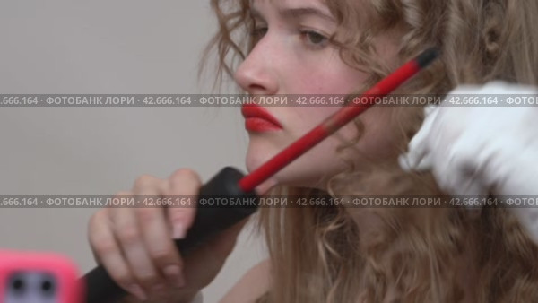 The woman with the beautiful long blonde hair is using a curling wand to style her hair. She's recording a vlog on phone while manually wrapping hair around the wand (2024 год). Редакционное видео, видеограф А. А. Пирагис / Фотобанк Лори