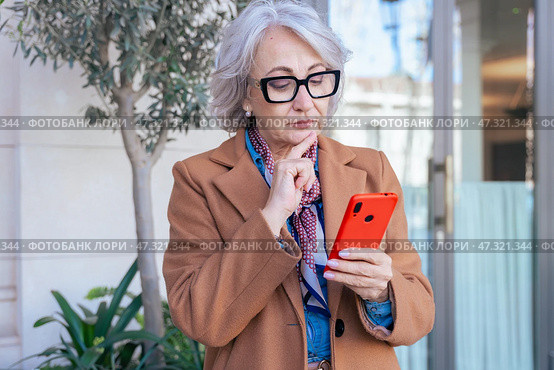 Thoughtful senior woman with smartphone looking away in an urban environment. Стоковое фото, фотограф Raquel Corcoles Mendez / Фотобанк Лори