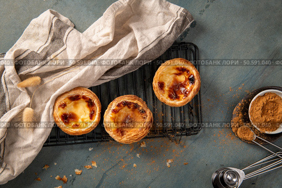 Three freshly baked Portuguese custard tarts on a cooling rack with a linen cloth and cinnamon ready to be sprinkled. Стоковое фото, агентство Ingram Publishing / Фотобанк Лори