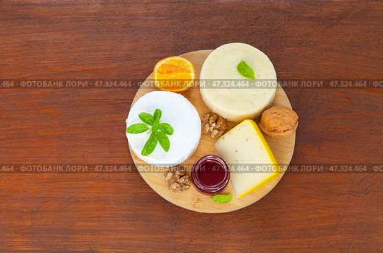 Three kinds of cream cheeses with basil, mandarin, fruit jam and walnuts on round cutting board on wooden table. Organic nutritious food. Top view, close-up, flat lay, copy space, mock up. Стоковое фото, фотограф Виктория Катьянова / Фотобанк Лори