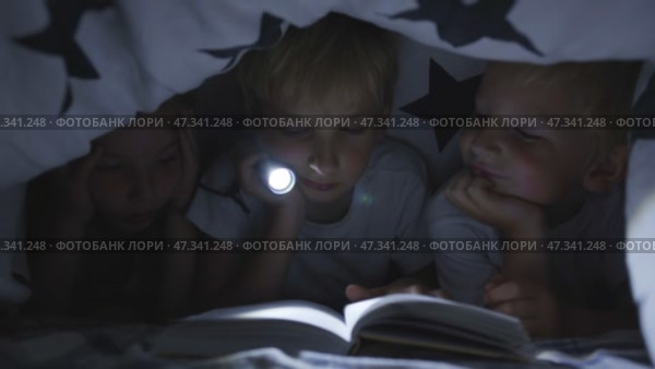 Three young children are reading a book with a flashlight under the covers at night. Стоковое видео, видеограф Mikhail Davidovich / Фотобанк Лори