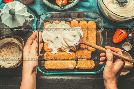 Tiramisu making.  Dish with italian savoiardi cookies and tiramisu ingredients on rustic kitchen table, top view. Woman hands holding a spoon and smeared cream on cookies.  Italian life style. Стоковое фото, агентство Ingram Publishing / Фотобанк Лори