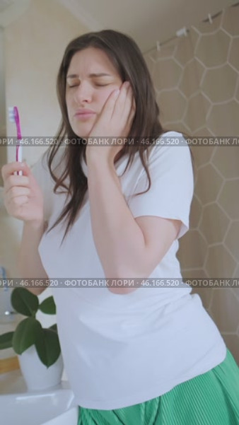 Tooth pain makes her hold her cheek, but oral care is important. Girl brushes her teeth in the bathroom despite cheek pain and discomfort. Стоковое видео, видеограф Svetlana Golovco / Фотобанк Лори