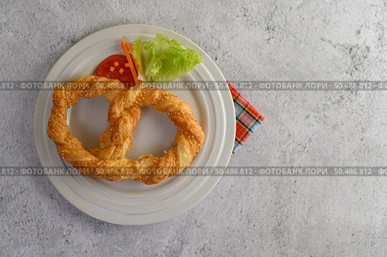 Top View Freshly baked soft pretzel with generous sprinkling of coarse salt and sesame on white dish, decorate with tomato sliced, lettuce and carrot beautifully, copy space. Стоковое фото, агентство Ingram Publishing / Фотобанк Лори