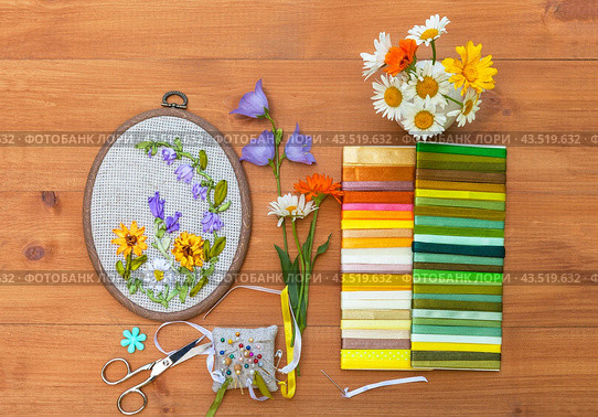 Top view of floral hand embroidery with satin ribbons of summer flowers in hoop and accessories for embroidery. Bouquet of daisies, calendula and blue bells on wooden background. Flat lay, close-up. Стоковое фото, фотограф Виктория Катьянова / Фотобанк Лори