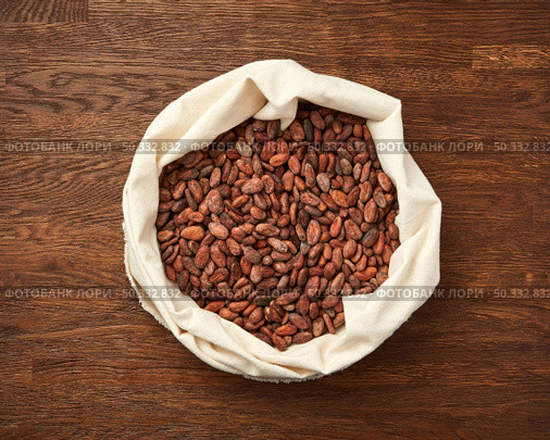Top view of textile white bag filled with raw unpeeled cocoa beans placed on rustic wooden surface. Sack of organic unpeeled cocoa beans. Стоковое фото, агентство Ingram Publishing / Фотобанк Лори