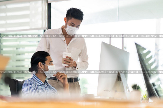 Two asian businessman talk discuss their work in morning after office reopen due to coronavirus COVID-19 pandemic. They wear protective face mask to prevent infection. New normal office life concept. Стоковое фото, агентство Ingram Publishing / Фотобанк Лори