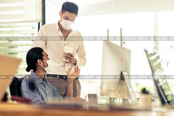 Two asian businessman talk discuss their work in morning after office reopen due to coronavirus COVID-19 pandemic. They wear protective face mask to prevent infection. New normal office life concept. Стоковое фото, агентство Ingram Publishing / Фотобанк Лори