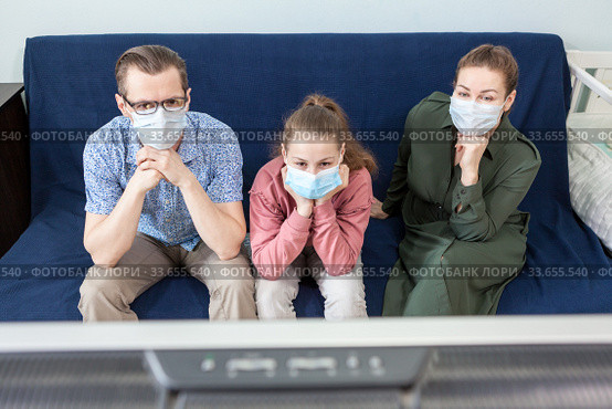 Two Caucasian parents with teenager daughter in medical masks sitting behind tv set on sofa in living room, people tired to watch television set. Стоковое фото, фотограф Кекяляйнен Андрей / Фотобанк Лори