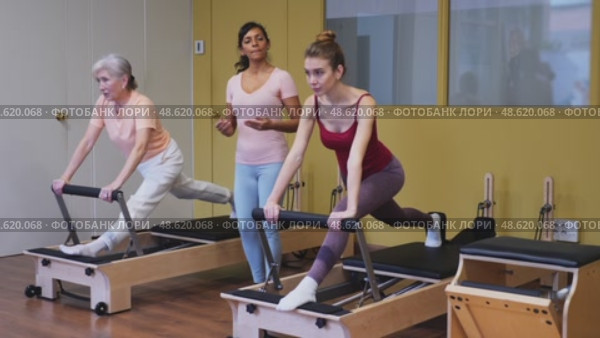 Two health conscious women of different ages perform a Pilates exercise using a reformer bed, where a female instructor helps them do it correctly. Стоковое видео, видеограф Яков Филимонов / Фотобанк Лори