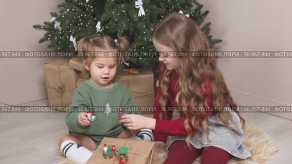 Two little girls, an older sister and a toddler, sit on the floor under the Christmas tree at home, playing with gifts and toys. Happy family and joy of winter holidays celebration concept. Footage. Стоковое видео, видеограф Ирина Михайличенко / Фотобанк Лори