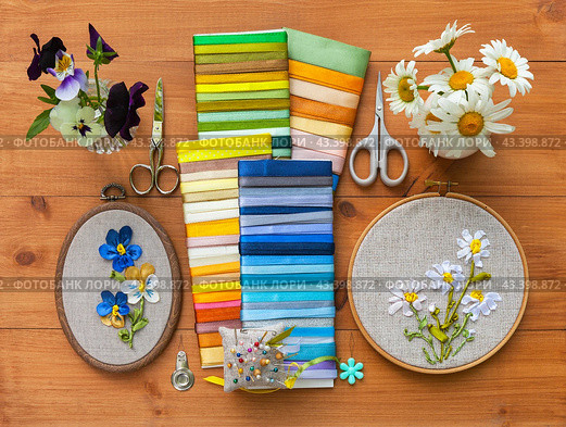 Two summer floral hand embroideries with satin ribbons of pansies and daisies in hoops, bouquets of flowers on wooden table. Nearby is set of satin ribbons, needles and scissors for work. Flat lay, close-up. Стоковое фото, фотограф Виктория Катьянова / Фотобанк Лори