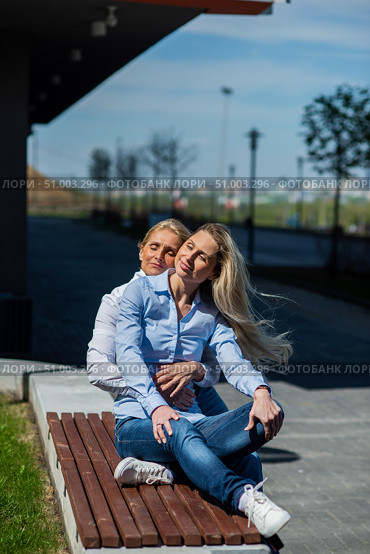 Two surprisingly similar women are sitting on a bench in an embrace and smiling. A middle-aged woman and her elderly mother. Beautiful well-groomed mother and daughter family look. Стоковое фото, фотограф Михаил Решетников / Фотобанк Лори
